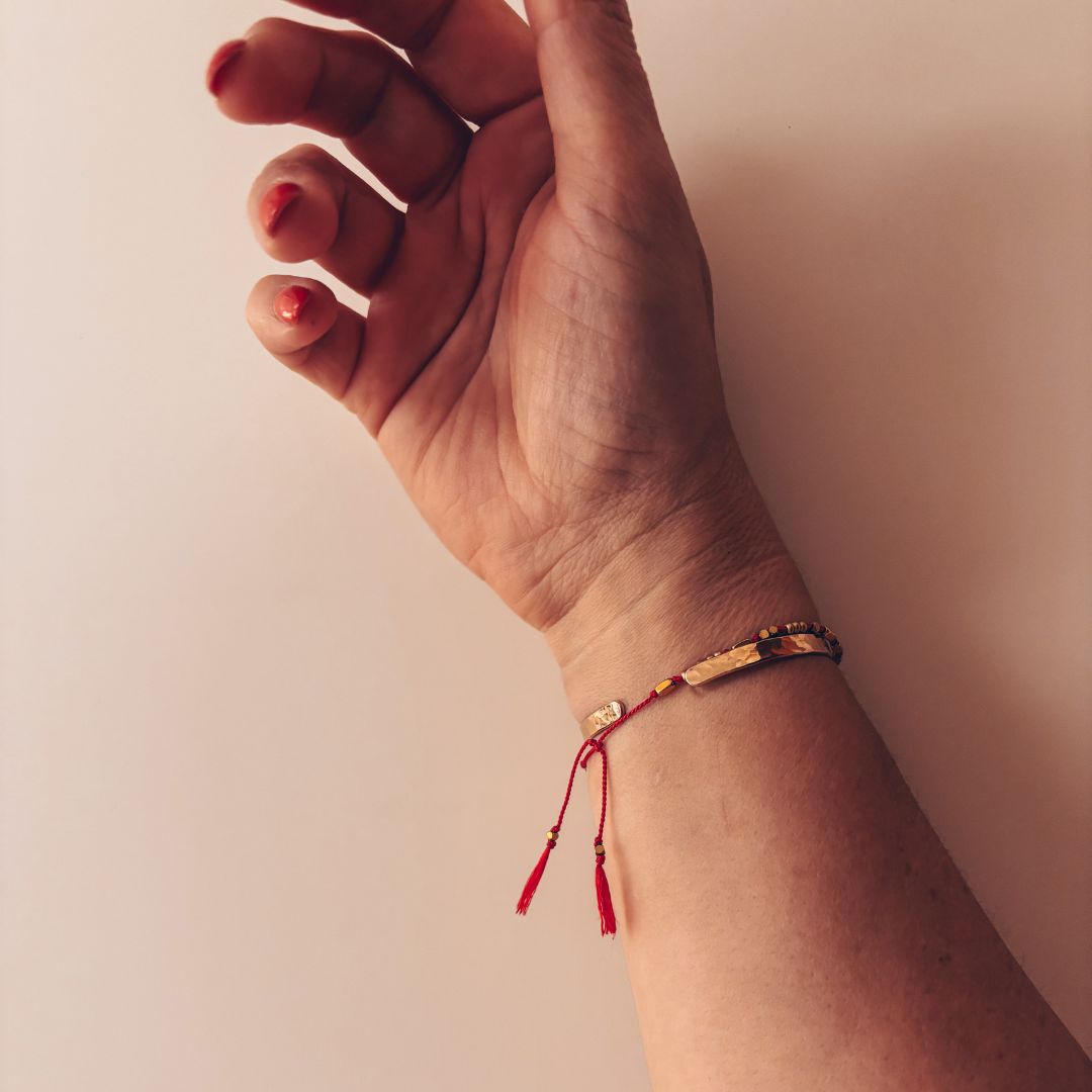 Bracelet fil rouge le Grigri MARLAH porté au poignet, perles dorées et pompons`