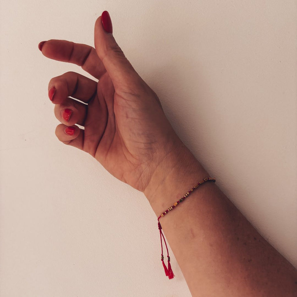 Bracelet fil rouge le Grigri MARLAH porté au poignet, perles dorées et pompons`